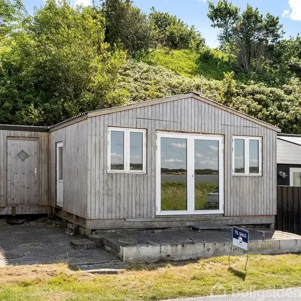 Et træhus står stille foran en bakke med buske. Et skilt ved vejen viser "Til salg". Huset har store vinduer og ligger ved en åben græsplæne.