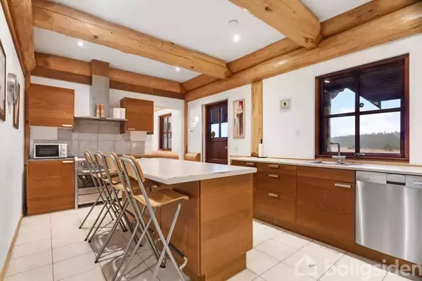 Køkken med træbjælker har et spisebord med høje stole. Køkkenskabe i træ er monteret, opvaskemaskine ved vindue. Lyst gulv og væg gør rummet indbydende.