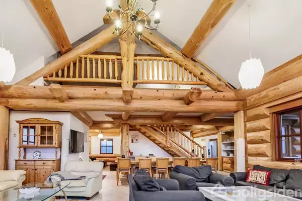 En stue med træbjælker dominerer, møbleret med sorte sofaer, spisebord og trappe til en hems. Omgivelserne inkluderer lysekroner og polstrede stole.