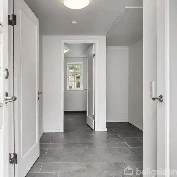 En indgangshal med grå flisegulv og hvide vægge fører til en døråbning. I baggrunden ses et vindue med udsigt til grønne træer udenfor.