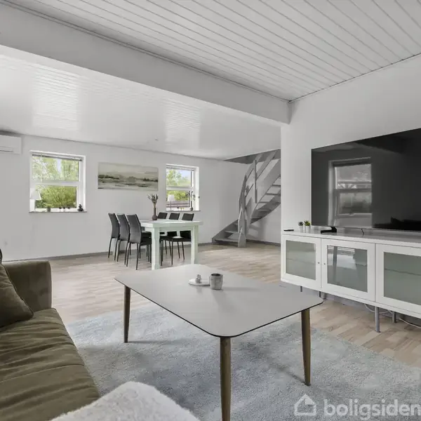 Stue med en stor sofa og sofabord foran et fladskærms-TV. Et spisebord med stole står nær vinduer. Trappe i baggrunden. Lys indretning med hvide vægge og gulv af træ.