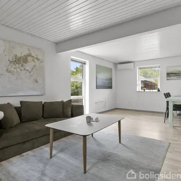 En stue med en brun sofa og et sofabord. På væggen hænger et verdenskort. Rummet har lyse trælofter, store vinduer, og i baggrunden ses et spisebord med stole.