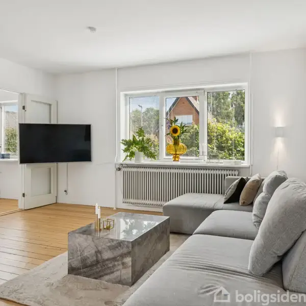 Grå sofa står i lys stue ved vindue. Et kvadratisk marmorbord har to stearinlys på. Stor fladskærm hænger på væggen. Sollys og planter skaber en indbydende atmosfære.