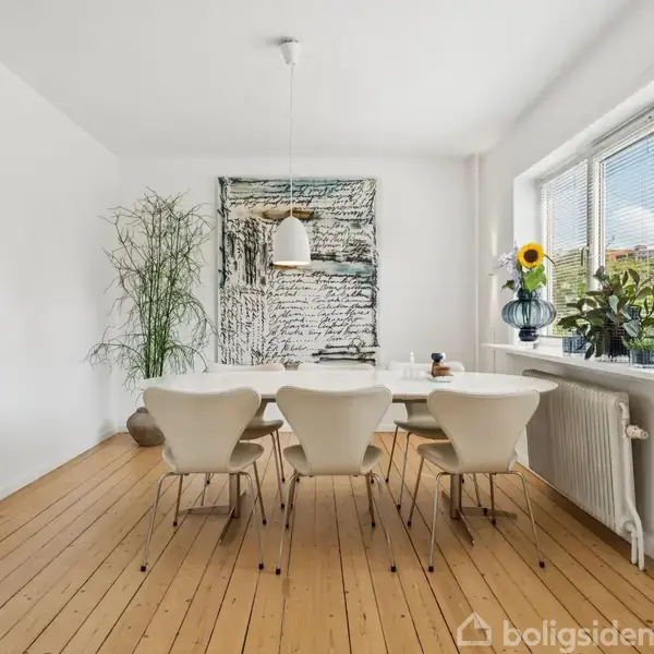 Fem stole står omkring et spisebord i et lyst rum med trægulve. Et kunstværk hænger på væggen, og planter pynter på en vindueskarm med udsigt til blå himmel.