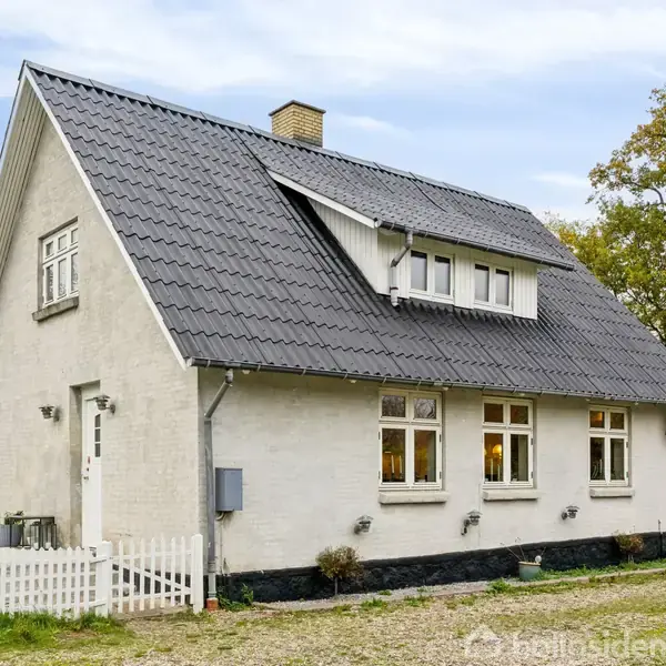 Et hvidt, klassisk hus med sort tegltag står ved siden af en grusvej omgivet af grønne træer og en lille hvid stakit. En stor græsplæne ses i baggrunden.