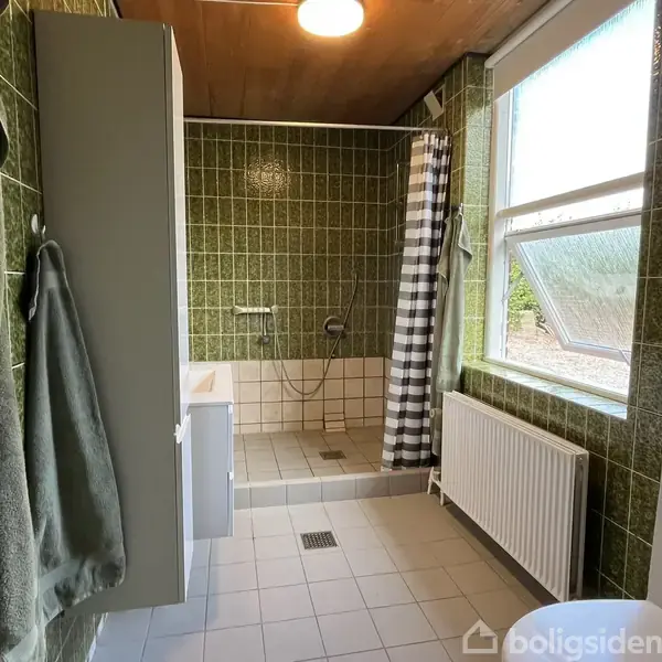 Brusekabine med grønne fliser og et stribet forhæng står i et badeværelse. En vindue med halvåben rude lader lys ind. Radiator og håndklæder hænger til venstre.