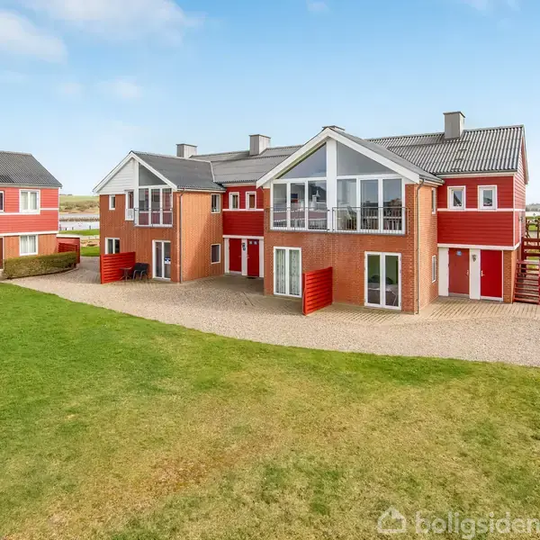 Rødt rækkehus med hvide detaljer og store vinduer står stille. Beliggende i et åbent, grønt område med græsplæne og blå himmel. Flere ens huse ses i baggrunden.