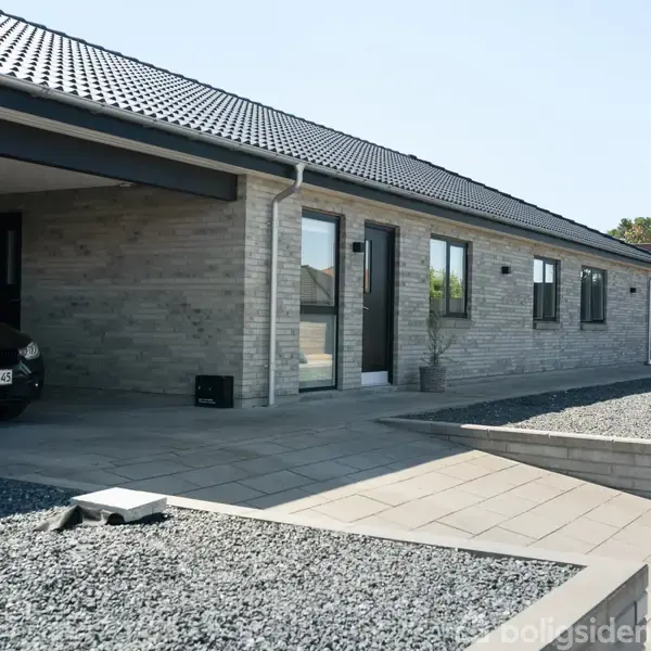 Et moderne murstenshus med sorte døre og vinduer. Foran huset er der en flisebelagt terrasse og indkørsel kantet med grus. En bil er delvist synlig under en carport.