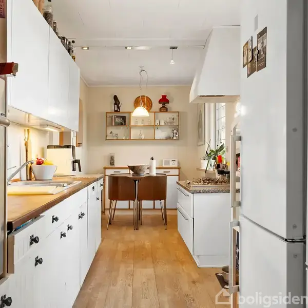 Et hvidt køkken med træbordplader, hvor ovne og skabslåger er synlige. Bordet er pyntet med en frugtskål. I baggrunden ses et spisebord med læderstole og en moderne lampe.