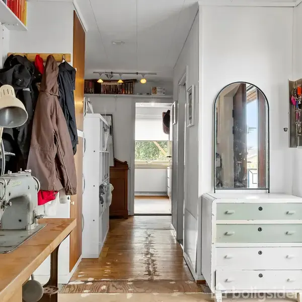 Symaskine står på et træbord til venstre, i en entré med jakker hængende på knager. Til højre ses en hvid kommode med spejl, og i baggrunden ses et lyst rum.