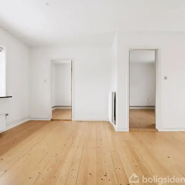 Et lyst rum med trægulv har to åbne døråbninger i baggrunden. Et vindue til venstre slipper naturligt lys ind, og væggene er hvide.