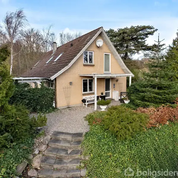 Et gult hus står stille omgivet af en frodig have med buske og træer, og en stentrappe fører op til indgangen.