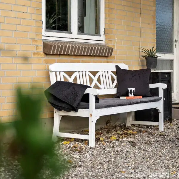 Bænk med sorte puder og et tæppe står foran et murstenshus ved indgangen, omkranset af grus og en plante i en potte. Det skaber en hyggelig indgangsidyl.