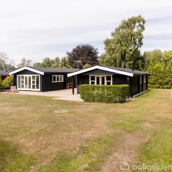 Mørkt træhus står stille i en velholdt have med græsplæne og enkelte buske. Huset har store vinduer, en terrasse, og er omgivet af grønne træer, hvilket skaber en fredelig atmosfære.
