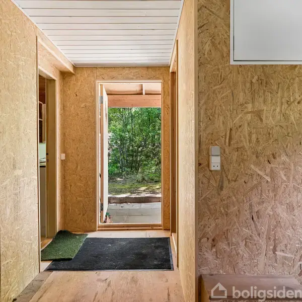 En gang i et hus med trævægge og træloft. En åben dør fører ud til en grøn have. En sort måtte ligger på gulvet ved indgangen.