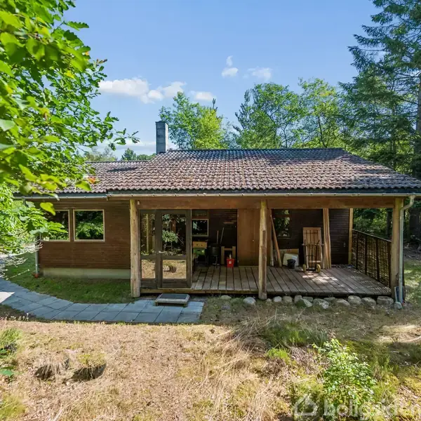 Et træhus med overdækket terrasse står stille i en frodig skovhave. Omkring huset ses der tæt bevoksning af træer og en græssti fører til indgangen.