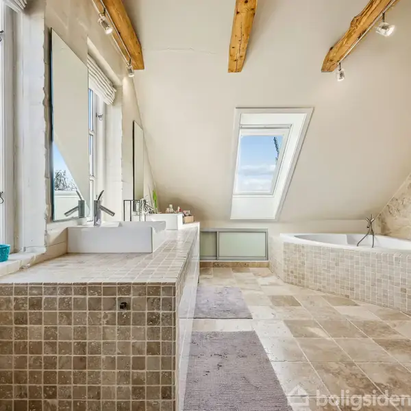 Badeværelse med skrånende loft og synlige træbjælker. Et badekar står ved væggen, og en stor håndvask med spejl ses ved vinduerne, der giver naturligt lys.