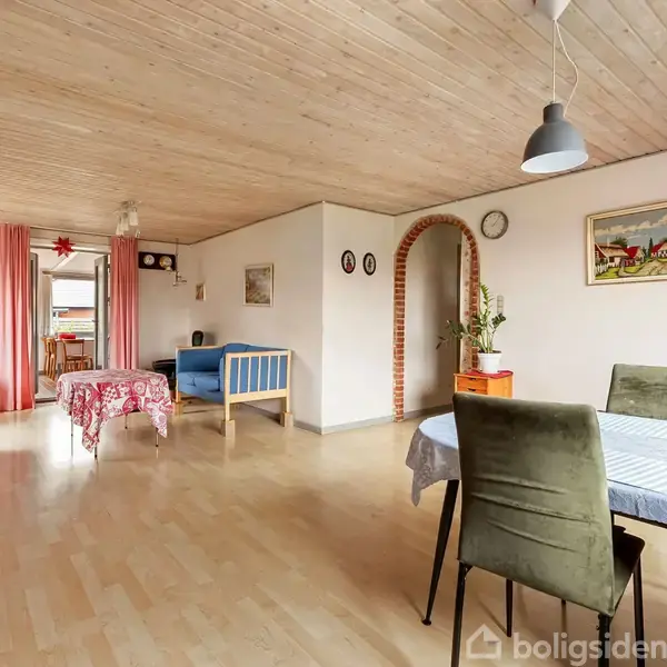 Rummeligt værelse med spisebord dækket af dug, omgivet af stole. I baggrunden står en sofa, reol og senge. Træloft og lyse trægulve skaber en hyggelig atmosfære.