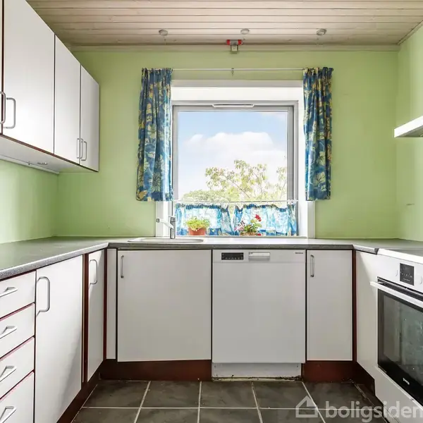 Et køkken med hvide skabe og gråt bordplade er i L-form. Der er et vindue med blå gardiner, og en opvaskemaskine står under det. Udenfor ses træer.