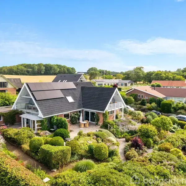 Et moderne hus med solpaneler sidder blandt velplejede haver med buske og blomster. Det ligger i et grønt kvarter med flere andre huse i baggrunden under en klar himmel.