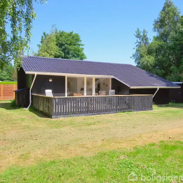 Et sort sommerhus med terrasse viser udendørs stole placeret foran huset, omgivet af en græsplæne og grønne træer i en skovlignende have.