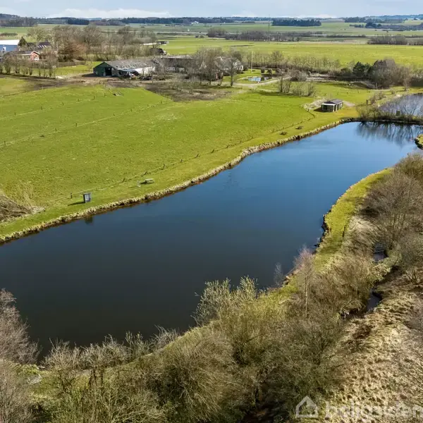 En stor, langstrakt dam ligger stille omgivet af grønne marker og træer. Et landligt landskab med en gård ses i baggrunden under en klar himmel.