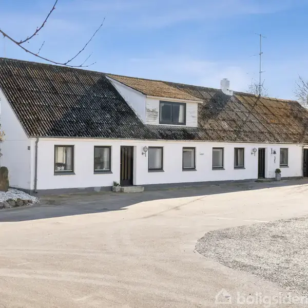 Hvidlakeret bygning med flere døre og vinduer, står på asfaltvej. Omkringliggende grusgård med få træer giver en landlig atmosfære.
