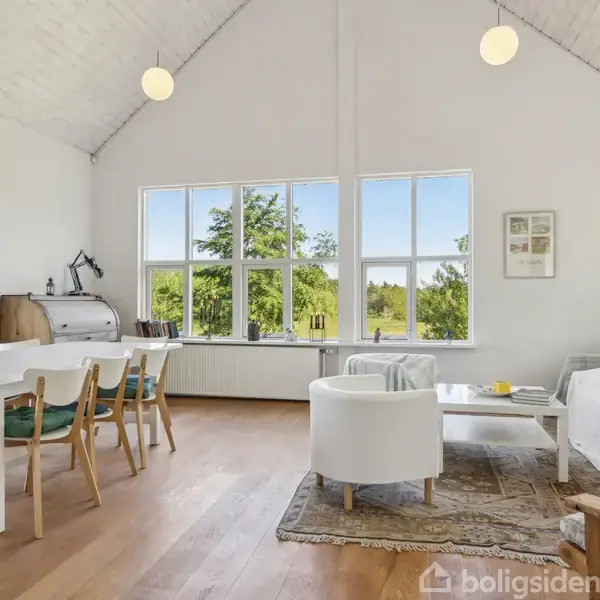Stue med loft til kip, indretning med hvidmalede vægge og træmøbler. En spisebord med seks stole og en sofa ved glasvægge. Store vinduer giver udsigt til grøn natur.