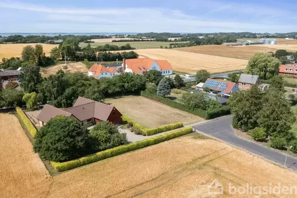 Luftfoto af en malerisk landsby med spredte bygninger, herunder huse med røde tage og marker omkring. Landskabet er frodigt og landligt, med veje der skærer igennem området.
