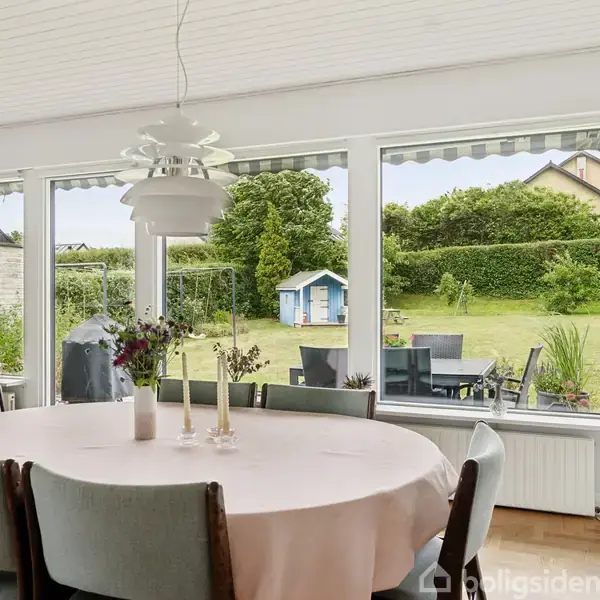 Rundt spisebord med lyserød dug og blomsterdekoration står foran store vinduer. Udenfor ses en velplejet have med en lille blå legehytte og grønne buske.