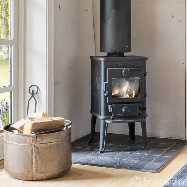 En brændeovn står på en flisebelagt sektion og er tændt med flammer. Til venstre står en spand med brænde. Rummet har en trædør, der fører ud til en terrasse.