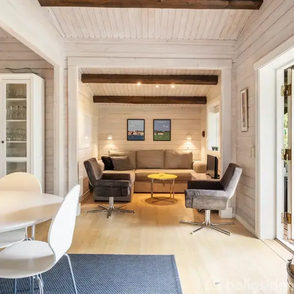Stue med grå sofa og to lænestole, rundt sofabord og spisebord med hvide stole. Store glasdøre åbner ud til terrasse med udsigt til have. Trævægge og loftsbjælker.