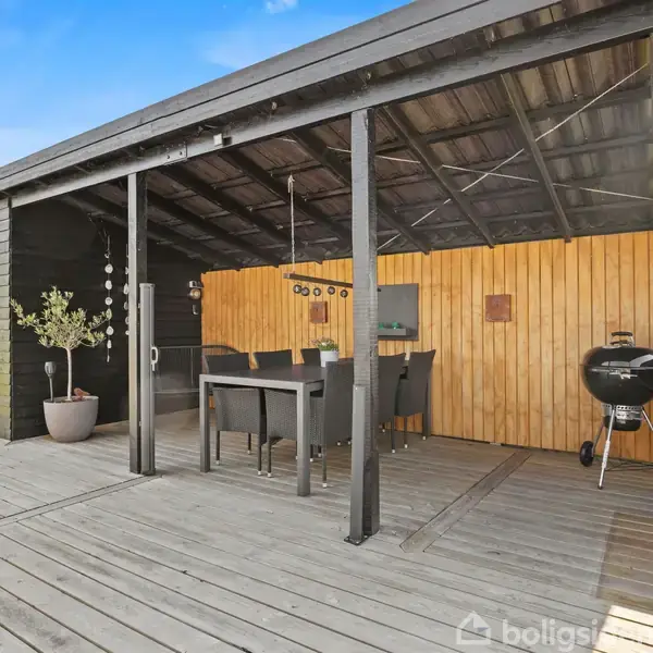 Overdækket træterrasse med sort bord og stole. Ved siden af står en kuglegrill. Baggrunden har vægge af lyst træ og en plante i krukke. Himmel er klar blå.
