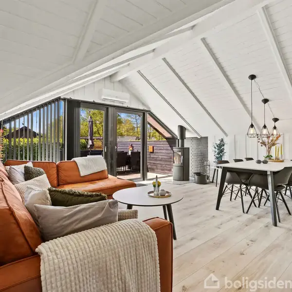 En stue med skrå lofter og hvide vægge. En brun sofa står til venstre, mens et spisebord med sorte stole er til højre. Store vinduer viser en terrasse udenfor.