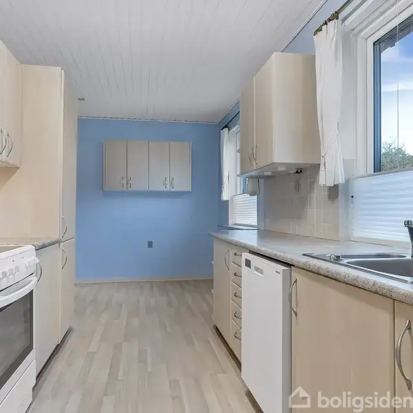 Køkken med hvid ovn og komfur til venstre; køkkenskabe og opvaskemaskine på begge sider. Et vindue med hvidt gardin kigger ud på grønne træer, blå vægge i baggrunden.