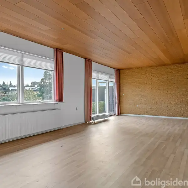 Et rummeligt, tomt værelse med trægulv, træloft og en murstensvæg. Store vinduer med røde gardiner giver udsigt til en have.