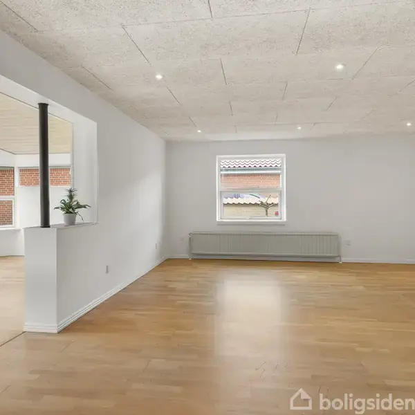 Stue med lyst trægulv beliggende i et rum med hvidmalede vægge. Et vindue viser udsigt til en murstensbygning. En åbning forbinder til et andet rum med planter.
