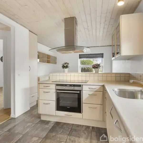 Et moderne køkken med lyse trækabelemøbler og en indbygget ovn. Køkkenet har flisegulv og vægfliser, en rund emhætte, og store vinduer med naturligt lys samt udsigt til planter udenfor.