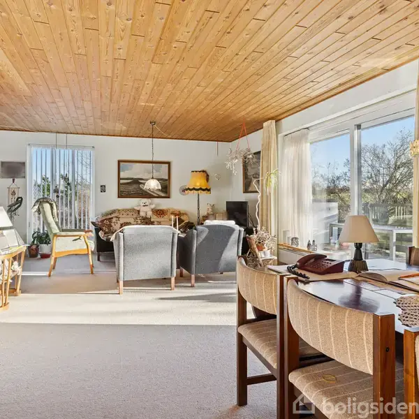 Stue med trægulv og -loft, hyggelige sofaer centreret om et sofabord, omgivet af store vinduer med haveudsigt. Et spisebord med stole er placeret i forgrunden.