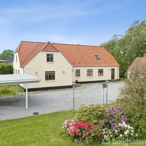 Hus med rødt tegltag og lyse vægge står ved en grusbelagt gårdsplads. Omkringliggende have har blomsterbed og buske. Carport ses til venstre, grønne træer i baggrunden.