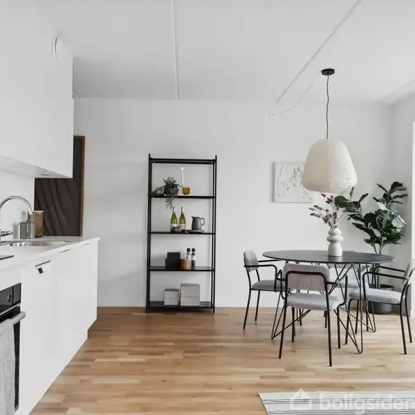 Et moderne køkken med hvide skabe, køkkenredskaber på bordpladen, og en bordplade i stål. Rundt spisebord med stole, reol med dekorationer, og en stor lampe hængende over spiseområdet.
