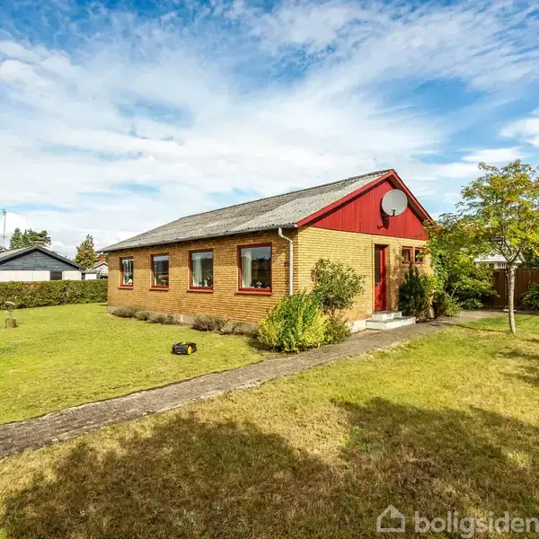 En gul murstensbungalow står på en velholdt græsplæne med få buske. Et dansk flag vajer på en stang i haven under en blå, let skyet himmel.