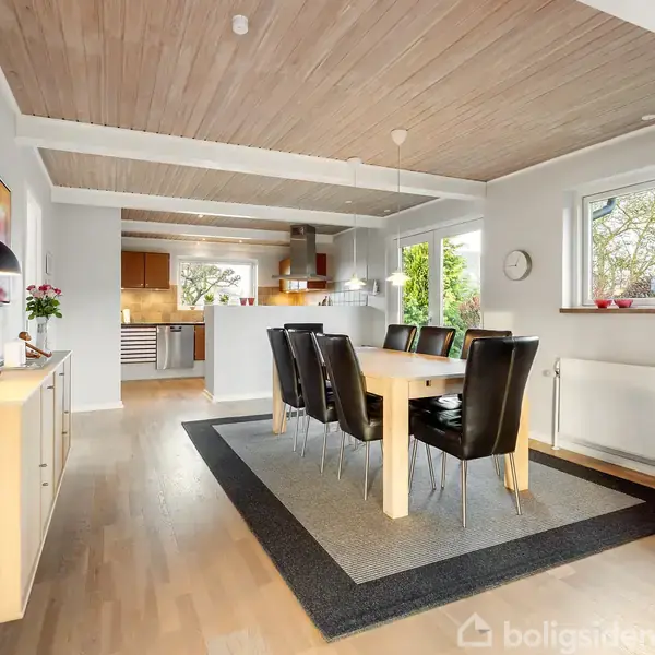 Et spisebord med sorte stole står på et tæppe i en moderne spisestue. Rummet har lyse trægulve, et træloft og store vinduer, som giver udsigt til en have.