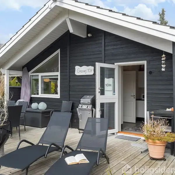 Sort træhus står på en træterrasse med liggestole, åben bog og grill. Dørskiltet viser "Granvej 10H". Terrasseborde og planter omkranser området, omgivet af træer og buske.