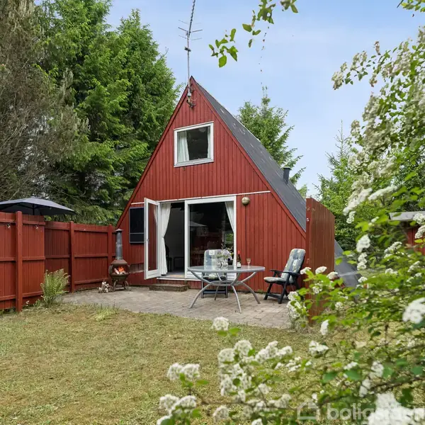 Rødt trehus med A-formet tag har åbne terrassedøre. To stole ved rundt bord på terrassen. Omgivet af rød træhegn og grønne træer. Blomsterbusk forrest.