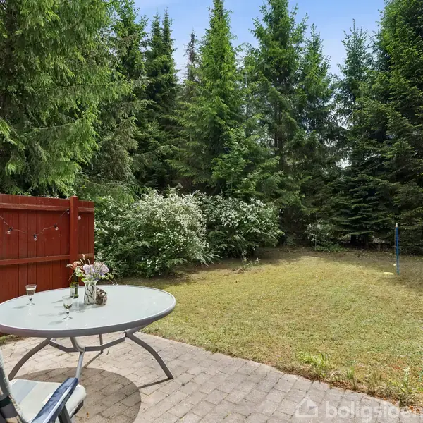Rundt bord med glas og vase står på flisebelagt terrasse. To stole ved bordet. Græsplæne og tæt bevoksning af høje grantræer i baggrunden. Ingen tekst til stede.