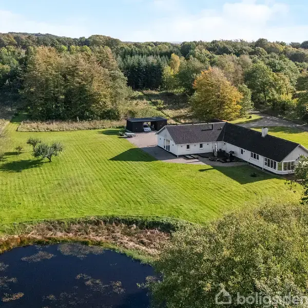 Et hvidt hus med sort tag ligger i en grøn have ved siden af en lille sø. Omgivet af skov og marker under en klar blå himmel.