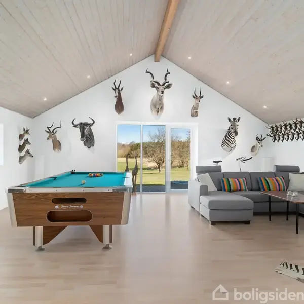 Et billiardbord står centralt i et rum med trofædyrhoveder på væggene. Til højre er en grå sofa med farvede puder. Gennemsigtige døre fører ud til en græsplæne. Poster med "Trans-Am" ses til venstre.