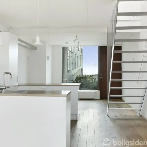 Køkken med hvide skabe og træbordplade, køkkenvask og ovn. En trappe fører til en øverste etage. Gennem vinduet ses en grøn udsigt og blå himmel.