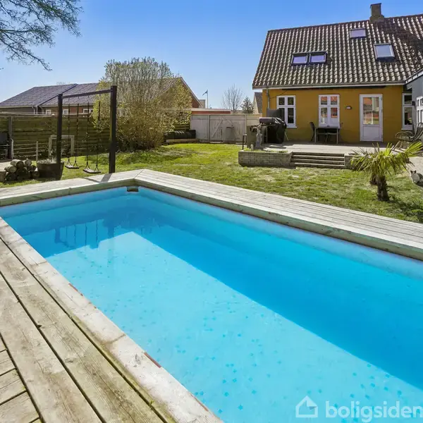Swimmingpool i en træterrasse omgivet af græsplæne og møbler, hvor et gult hus med mørkt tag står i baggrunden.